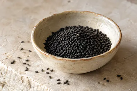 Graines de nigelle noires entières dans une coupelle en céramique posée sur une pierre claire.