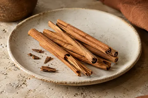 Bâtons fins de cannelle de Ceylan posés sur une céramique claire, avec quelques fragments près d’eux.