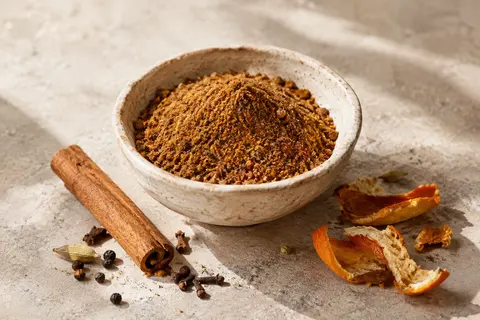 Mélange Baharat en poudre dans une coupelle de céramique sur une pierre claire, avec cannelle et écorces d’orange.