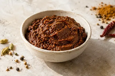 Pâte de curry Jalfrezi brun profond présentée dans une coupelle artisanale sur une pierre claire.