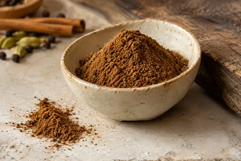 Poudre de garam masala brun chaud dans une coupelle en céramique posée sur une pierre claire.