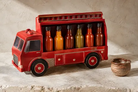 Coffret rouge en forme de camion de pompiers contenant six sauces piquantes, posé sur une pierre claire.